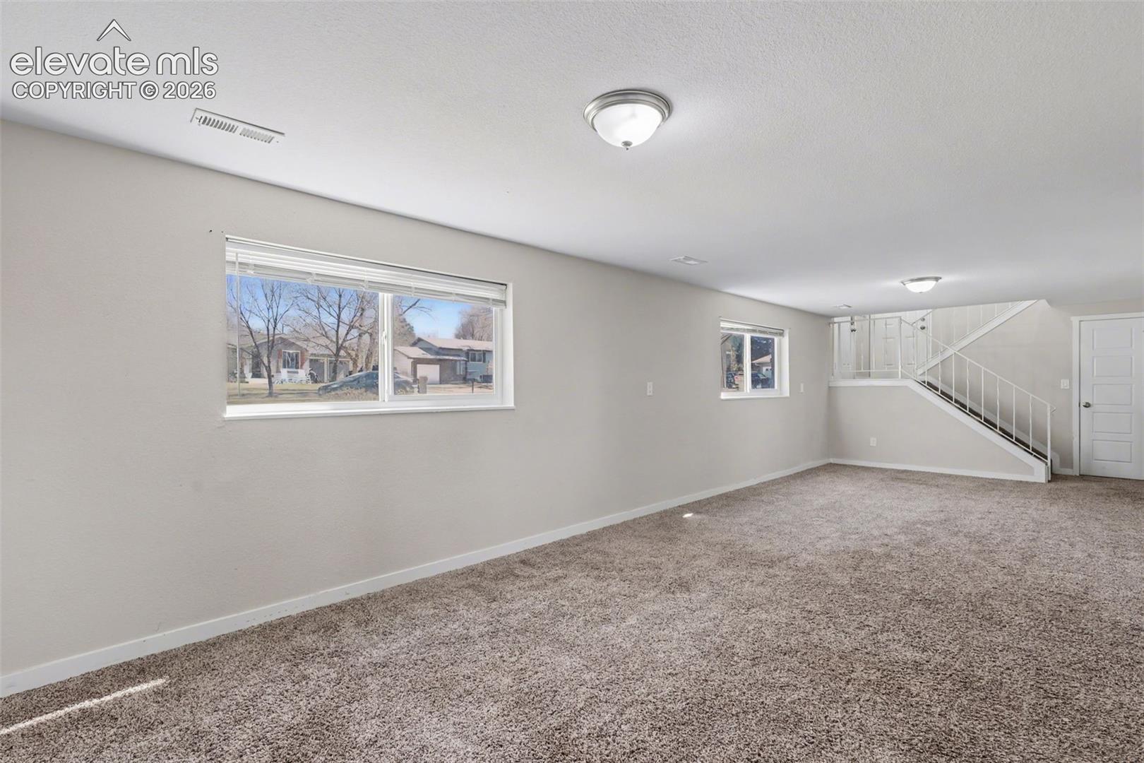 Neutral interior room featuring neutral wall paint, wall-to-wall carpeting, multiple double-pane windows, and a staircase with a metal railing