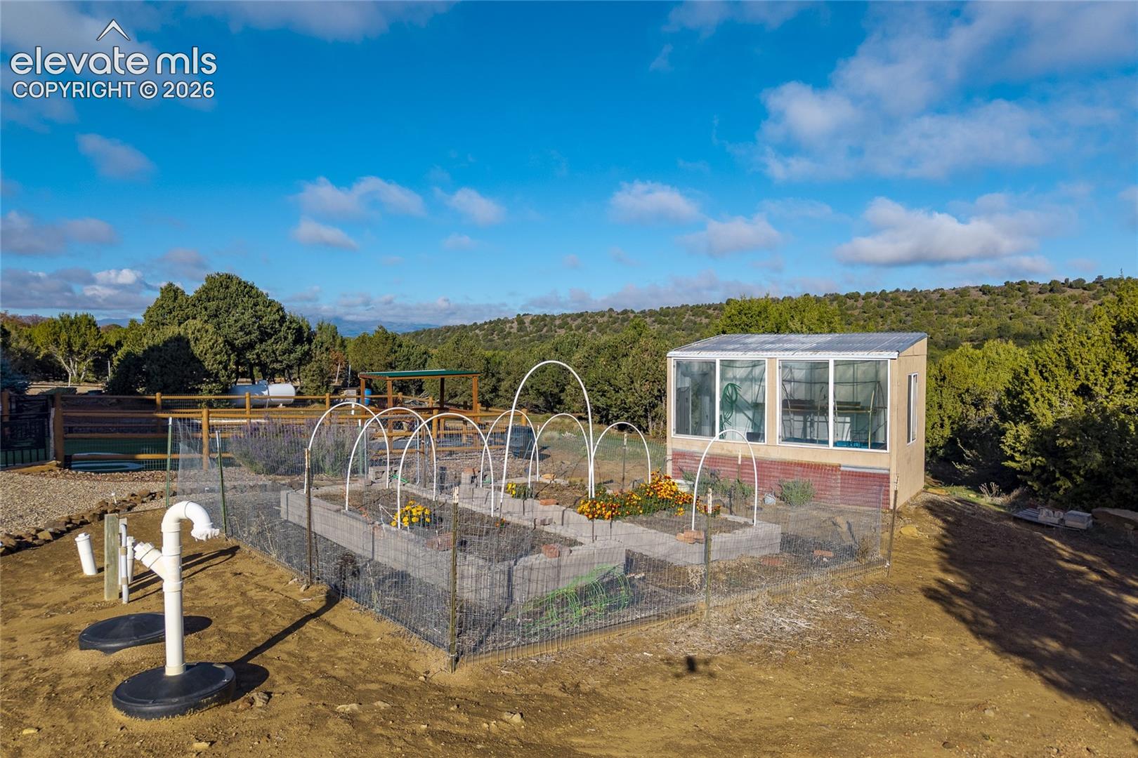 Enclosed garden area featuring masonry raised planting beds, metal hoop trellises, a standalone glass-paneled greenhouse, and an exterior water pump system