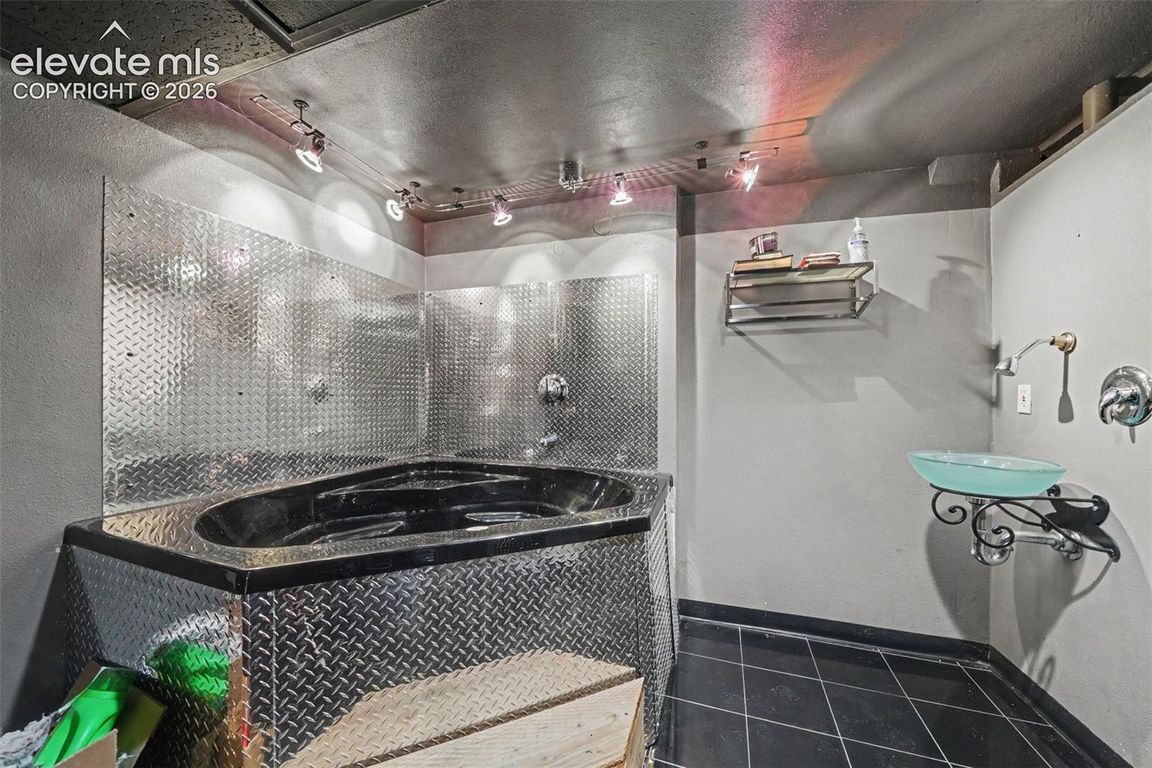 Corner soaking tub featuring a diamond-plate metal surround and wall panels, black tile flooring, wall-mounted glass vessel sink, and adjustable track lighting