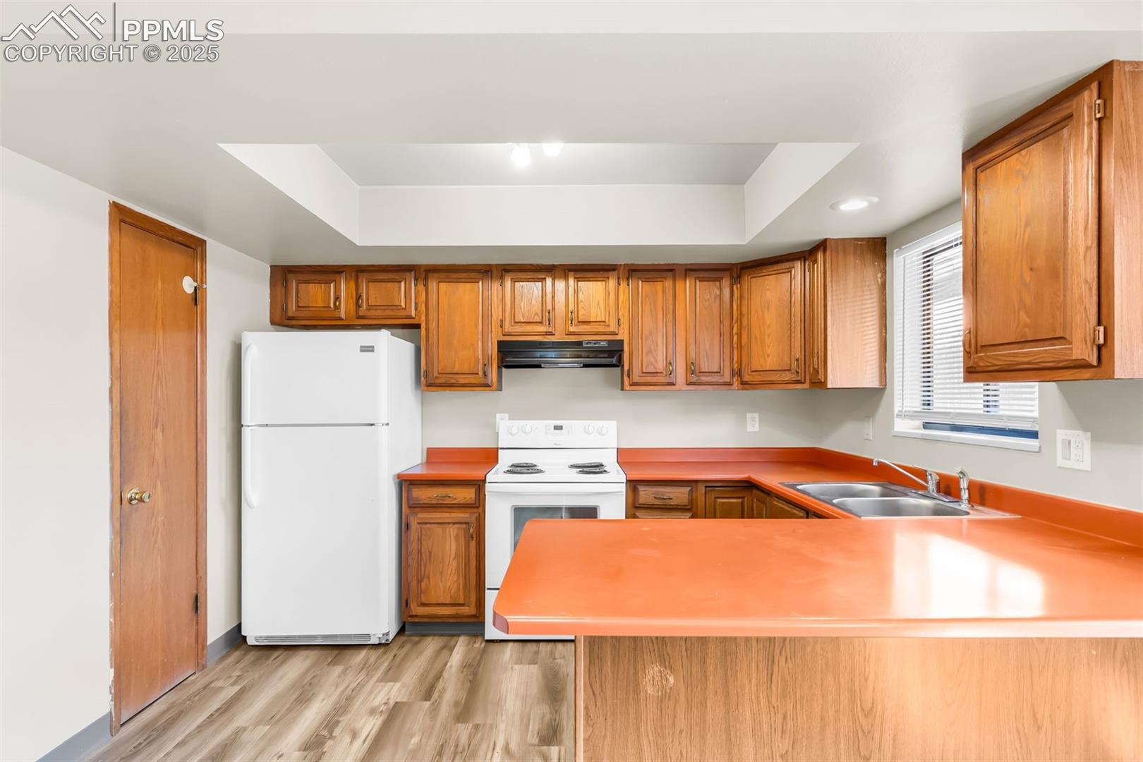 Kitchen with white appliances, light wood-type flooring, a raised ceiling, brown cabinetry, and recessed lighting