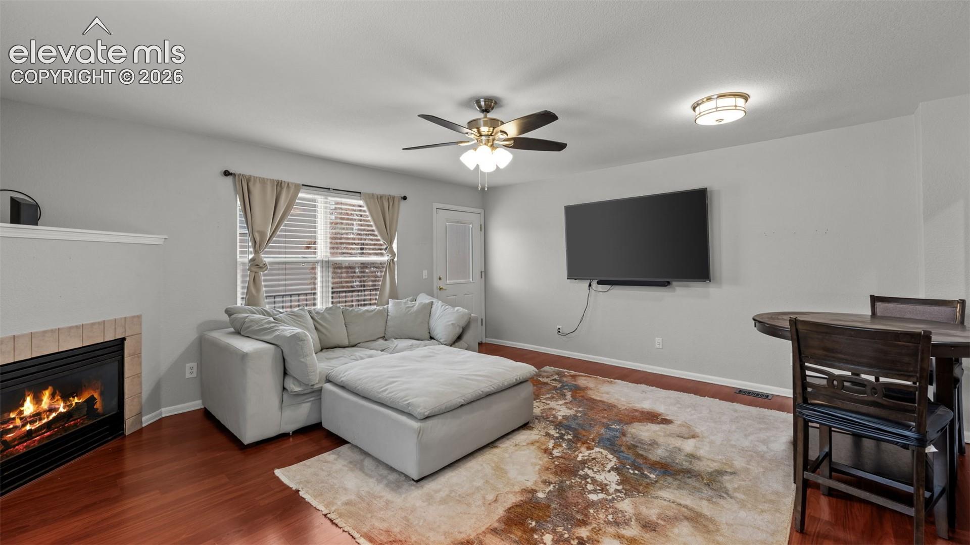 Fireplace with a tile surround, wood-finish flooring, ceiling fan with light kit, flush-mount lighting, and multi-pane window