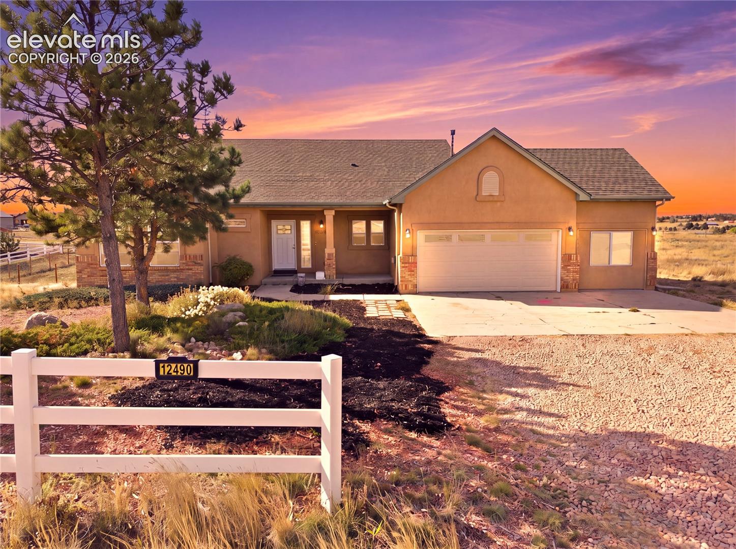 Stucco exterior featuring a dual-gabled roofline, stone veneer accents, an attached two-car garage, and a gated perimeter fence