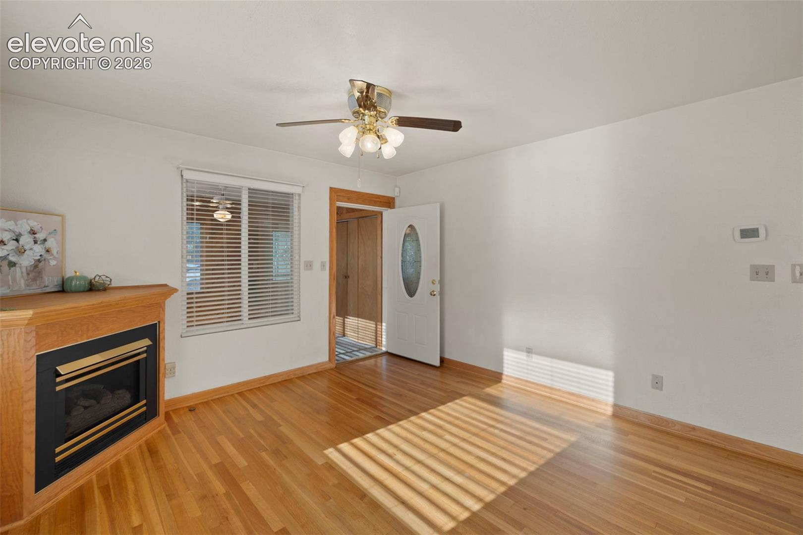 Wood-finish flooring, corner fireplace with a wood mantle, ceiling fan with integrated lighting, and a white entry door with an oval glass insert