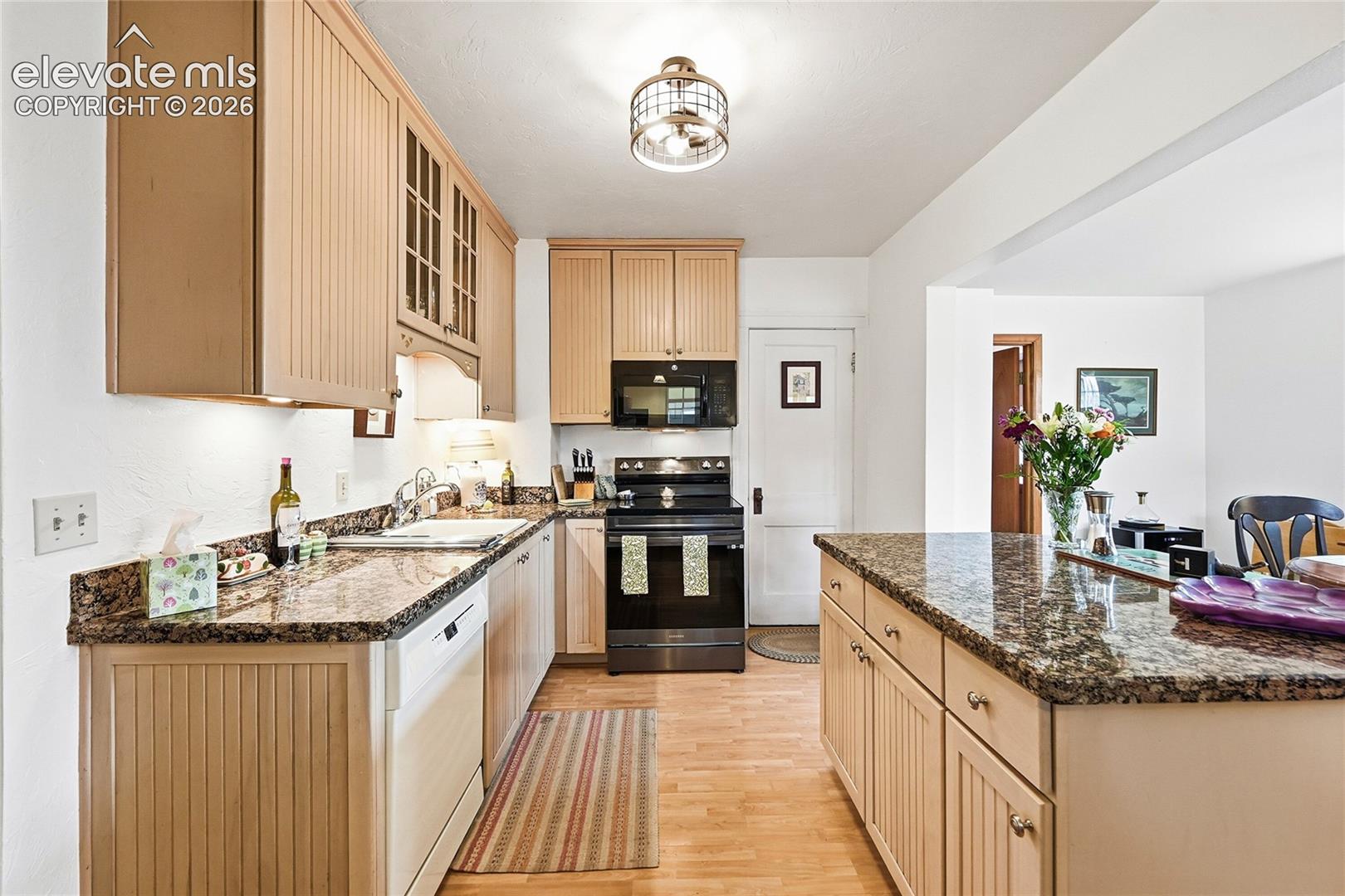 Kitchen featuring light wood cabinetry with glass-panel accents, stone countertops, black appliance suite, wood-finish flooring, and a drum-style pendant light fixture