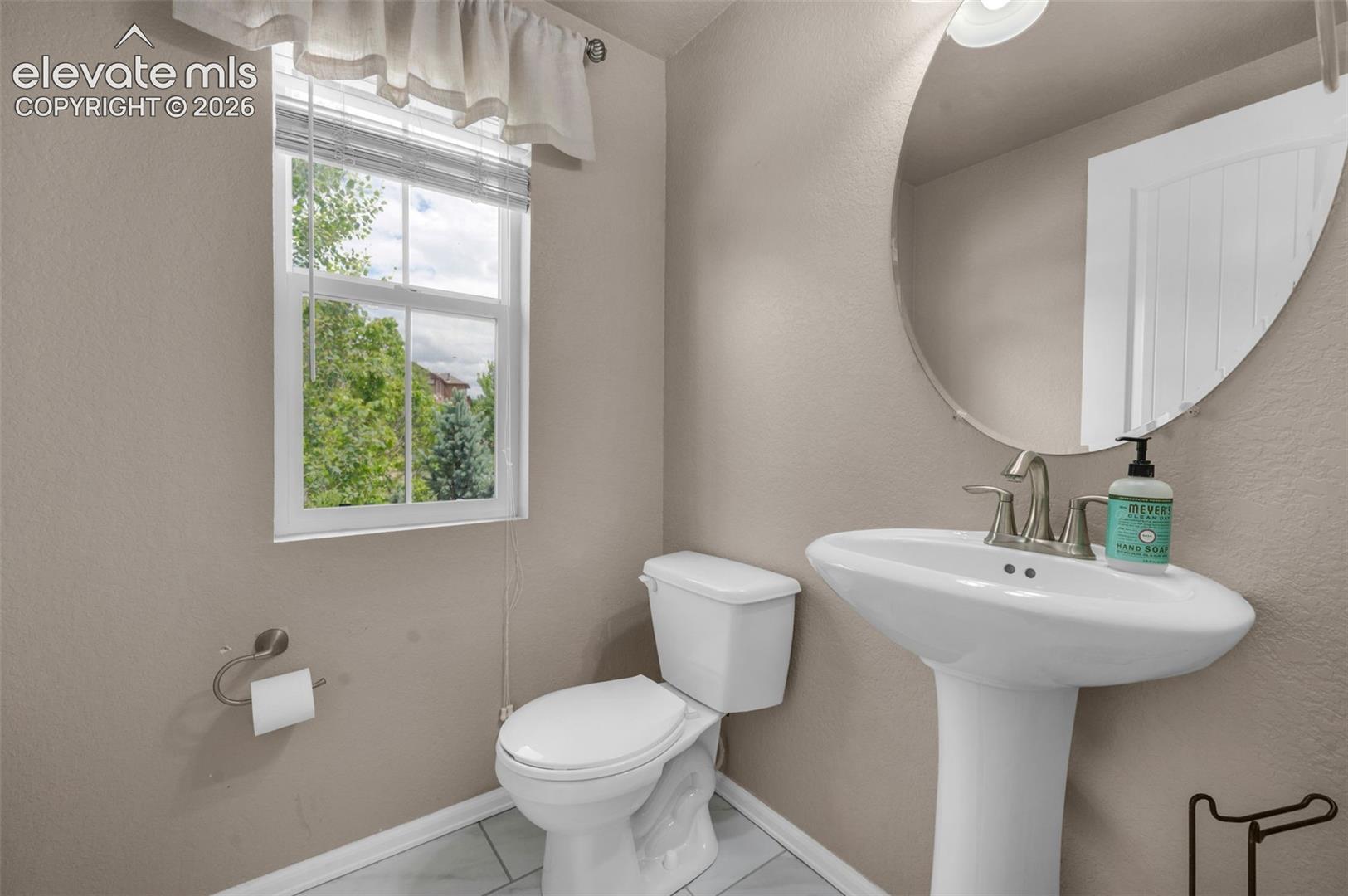 Powder room featuring a pedestal sink with brushed nickel faucet, a rounded wall mirror, and a window with blinds