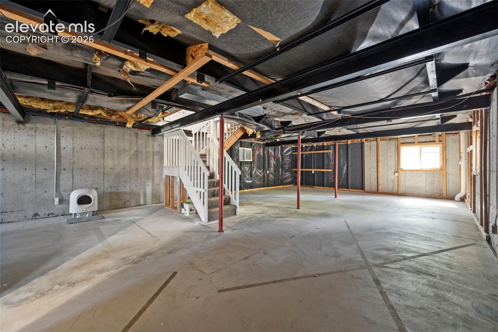 Unfinished lower level featuring concrete foundation walls, exposed steel structural beams, a concrete floor, and a wooden staircase with white balusters
