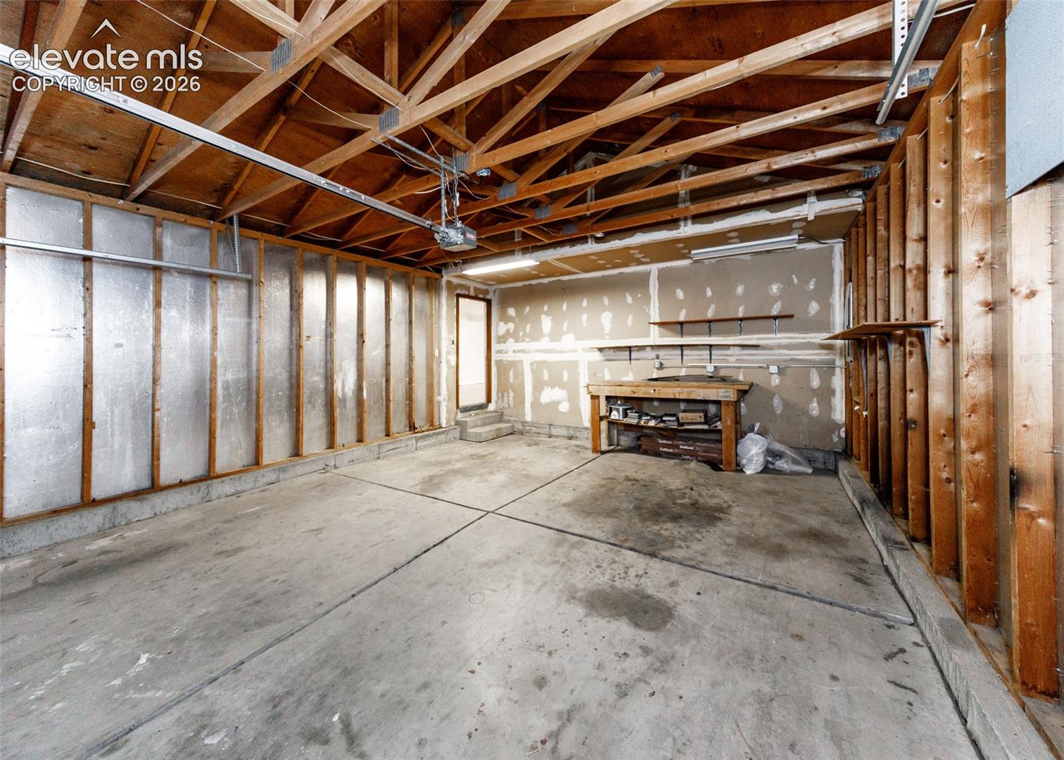 Garage space featuring exposed timber roof trusses, concrete flooring, reflective wall insulation, a built-in wooden workbench, and overhead fluorescent lighting