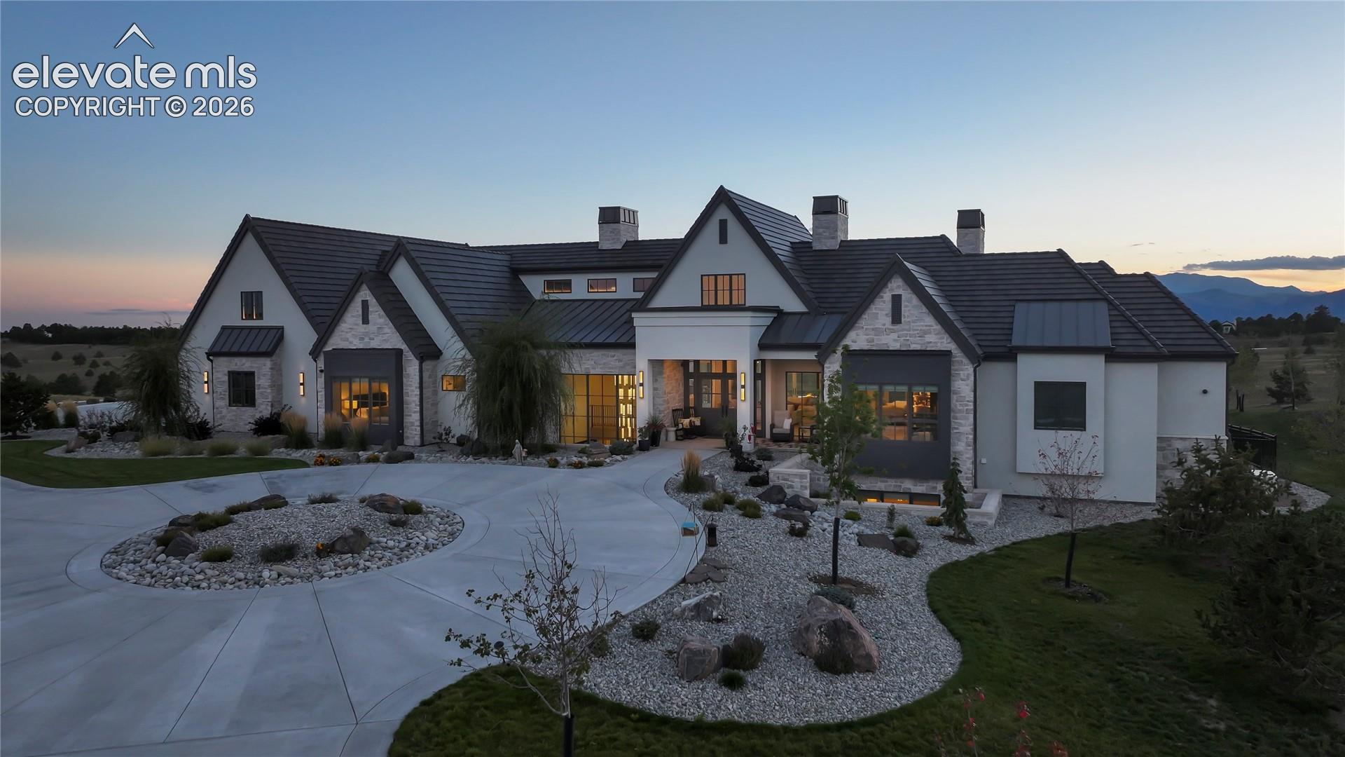 Custom estate featuring a stone and stucco facade, multi-gabled roofline, expansive circular concrete driveway, and professional xeriscaped grounds with decorative rock accents