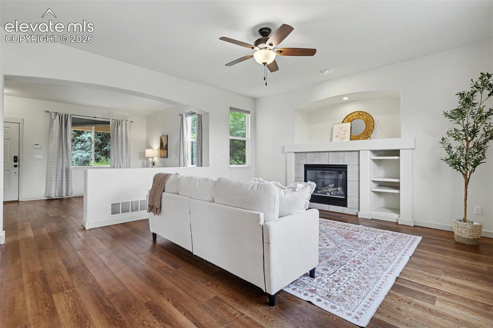 Living area featuring wood-finish flooring, a ceiling fan, and a fireplace with a tiled surround