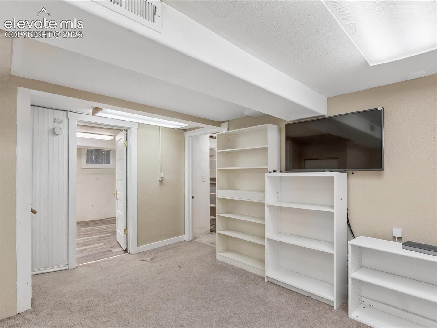 Carpeted room featuring exposed ceiling beams, a wall-mounted television, and multiple built-in shelving units