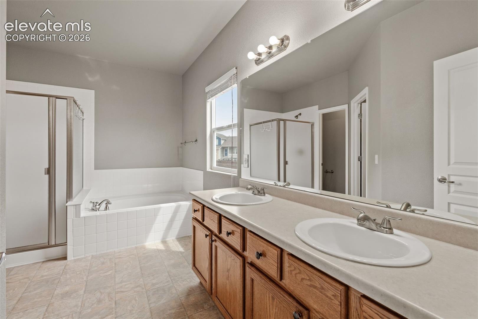 Bathroom featuring a dual-sink vanity with wood cabinetry, a tiled-surround soaking tub, a separate glass-enclosed shower, and a large wall-mounted mirror