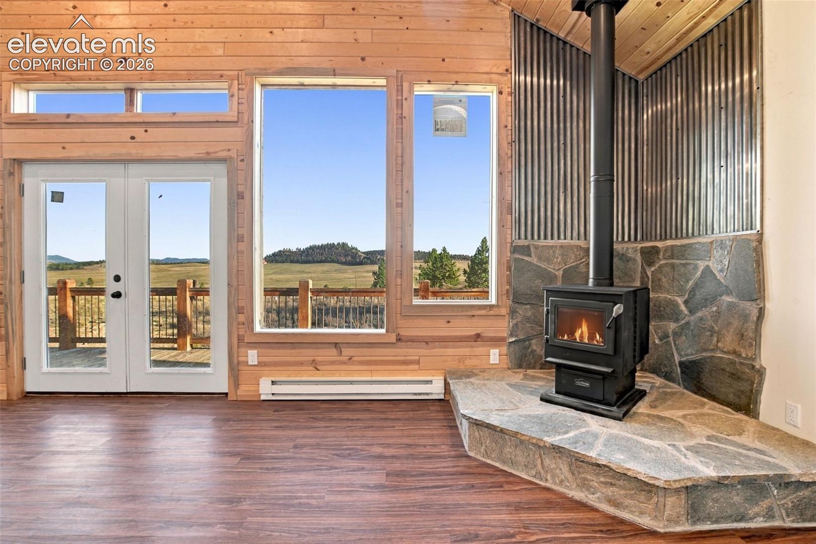 Wood-plank wall paneling, full-height windows, freestanding wood-burning stove with a stone hearth, corrugated metal accent wall, and wood-finish flooring