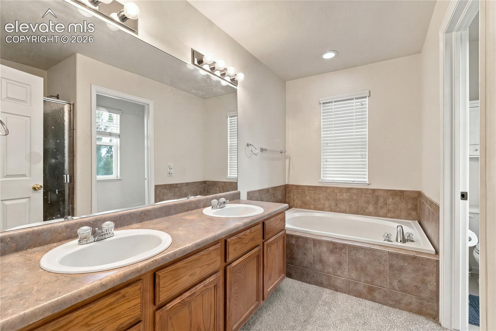 Double vanity featuring wood-finish cabinetry, solid-surface countertops, dual drop-in sinks, and a large wall-to-wall mirror, complemented by a tiled soaking tub