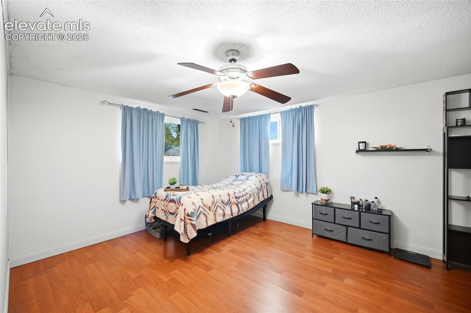 Wood-finish flooring, central ceiling fan with integrated lighting, dual windows, and wall-mounted shelving