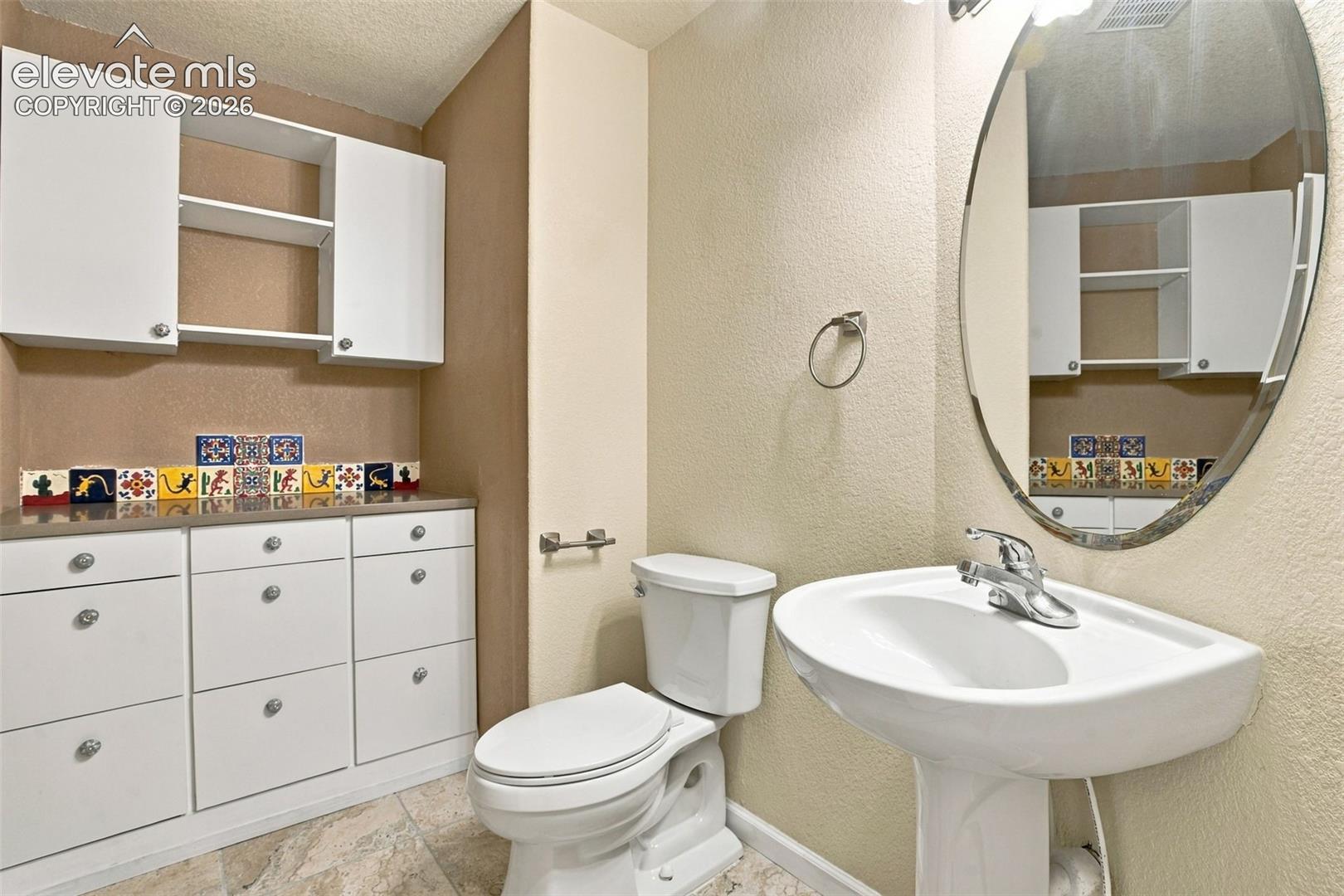 White built-in cabinetry with decorative tile backsplash, oval wall-mounted mirror, pedestal sink, and stone-finish tile flooring