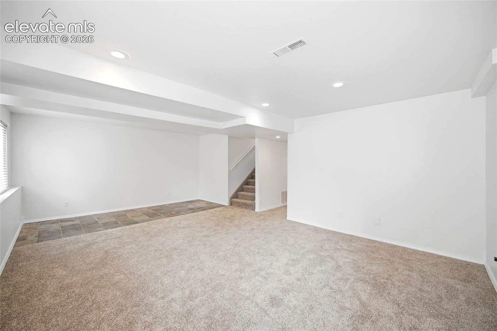 Neutral-toned interior space featuring recessed lighting, multi-textured flooring with carpeted and tile sections, and a staircase with wall-mounted railing