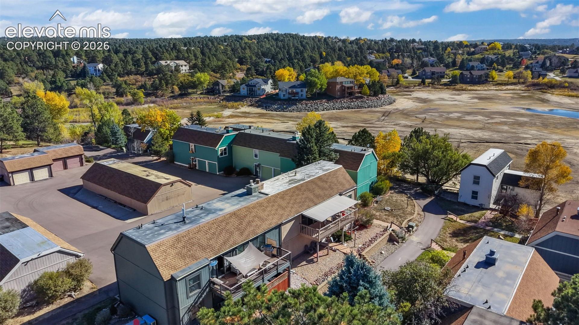 Aerial perspective of property featuring asphalt surface parking, multiple attached garage structures, wooden deck with railing, and mature pine tree landscaping