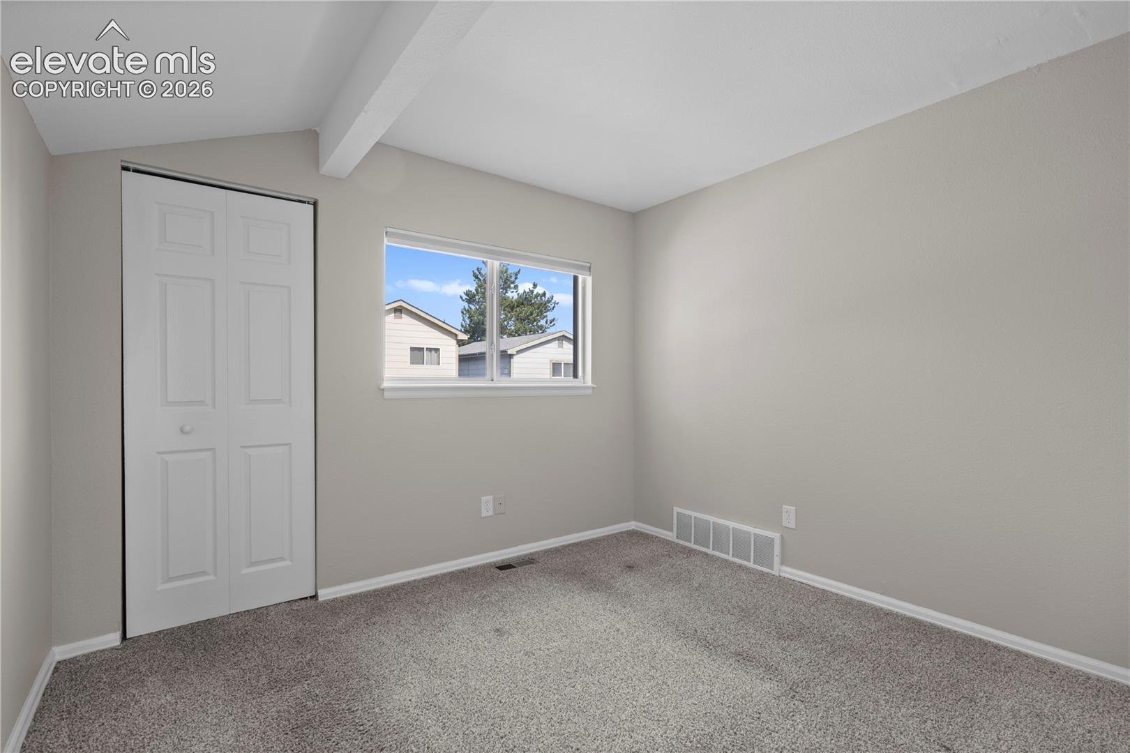 Neutral interior space featuring carpeted flooring, a vaulted ceiling with exposed structural beam, and a double-panel closet door