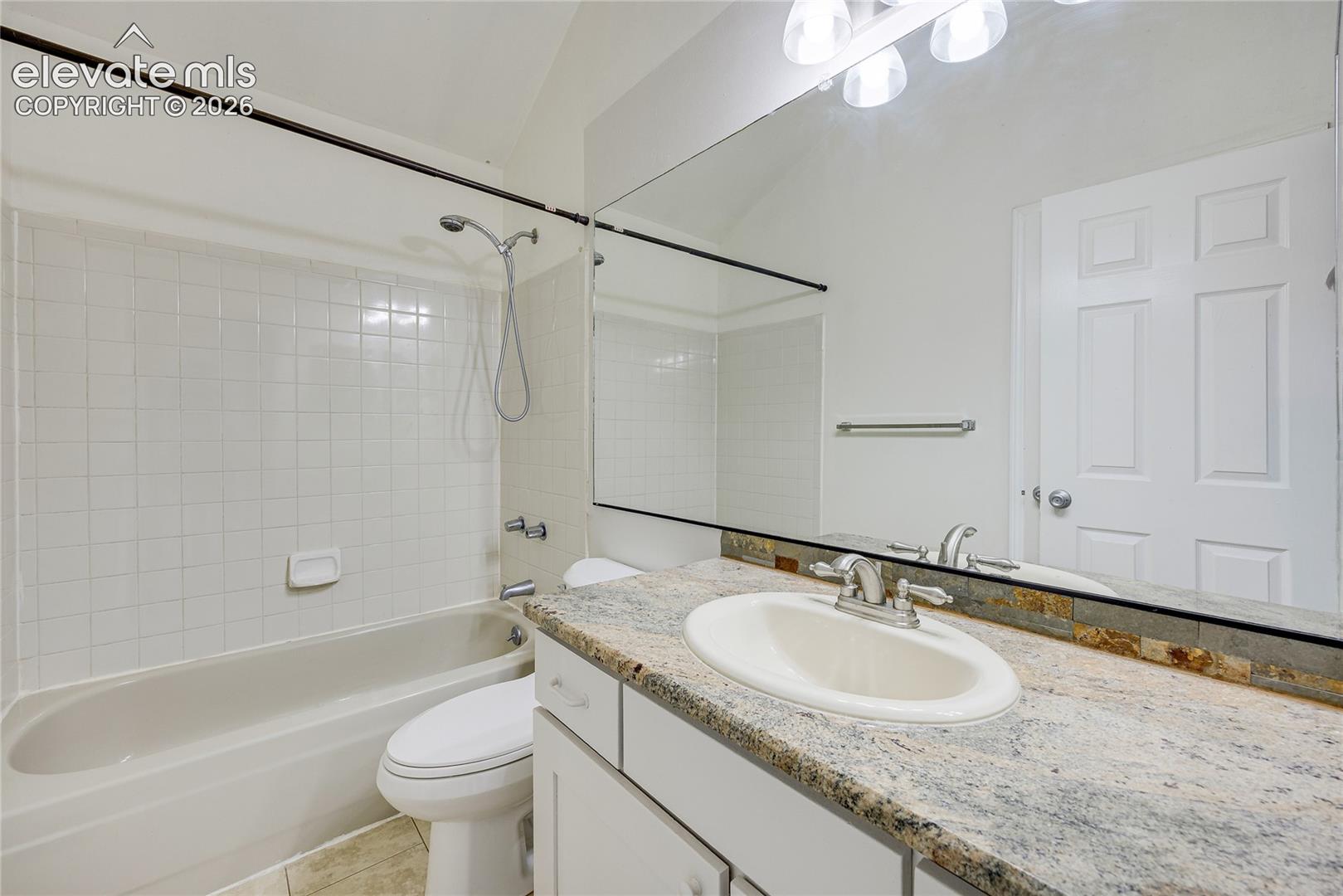 White rectangular tile shower surround, shower/tub combination, white vanity with integrated sink, granite-style countertop, and full-width mirror