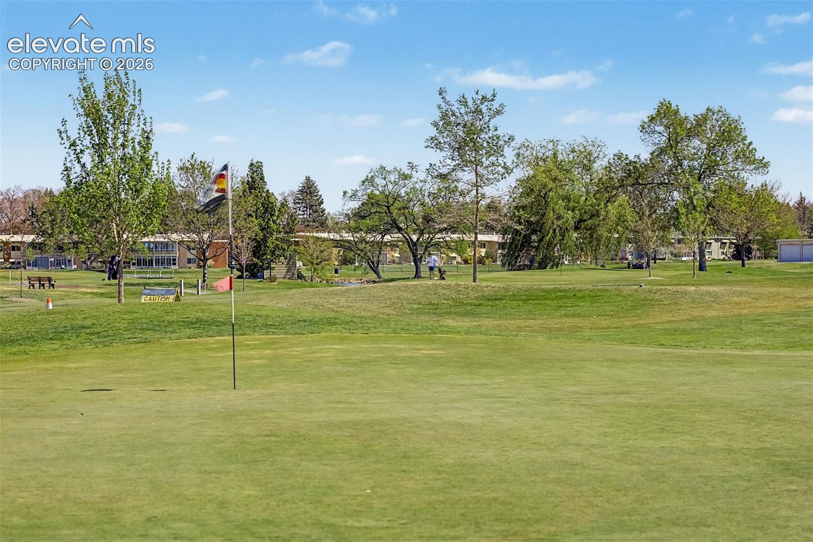 Expansive green golf course featuring a putting green with a flagstick, mature trees, and distant buildings