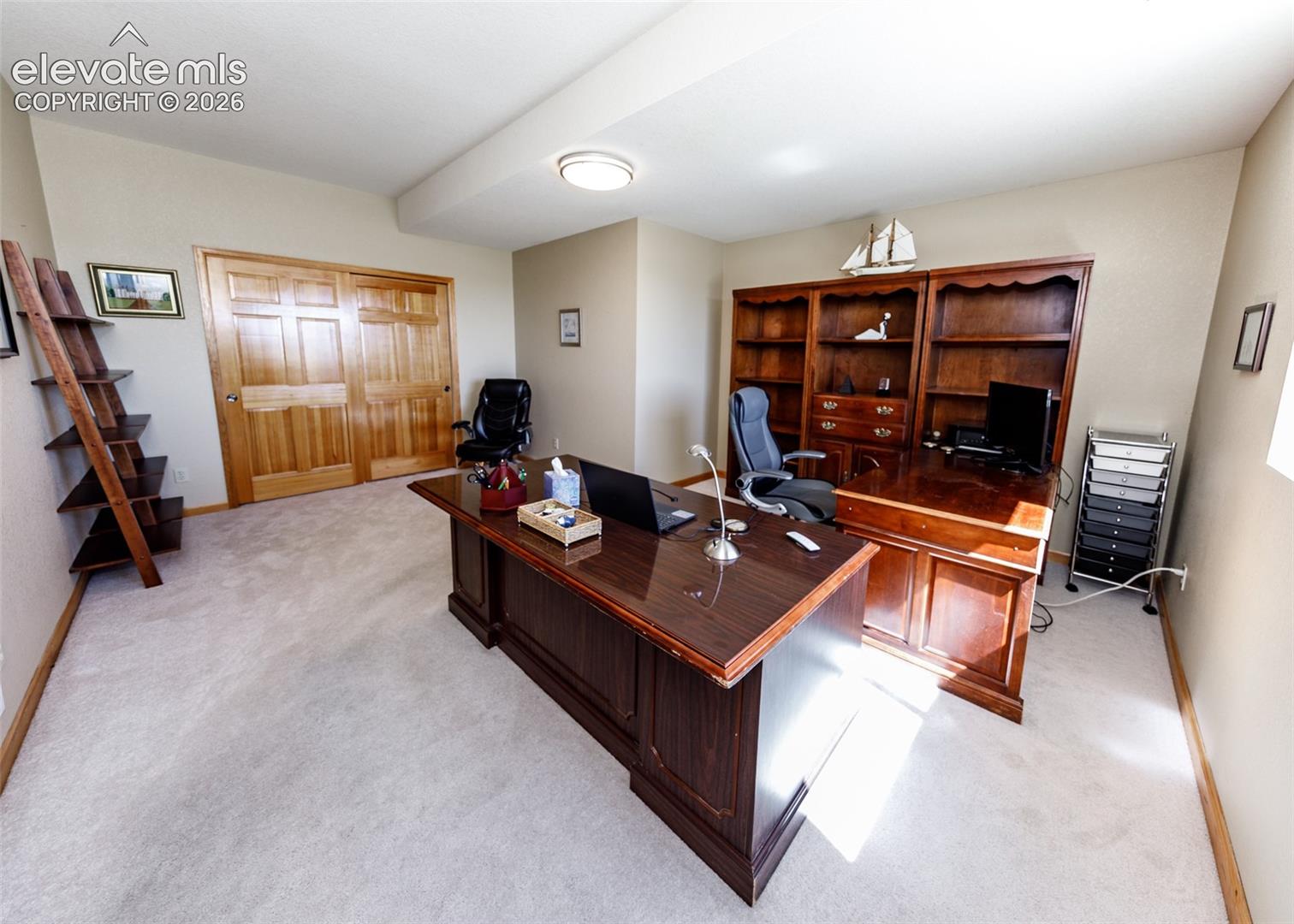 Sophisticated home office featuring a commanding wood executive desk, extensive built-in cabinetry with glass-front display shelving, recessed ceiling lighting, and neutral carpeting throughout