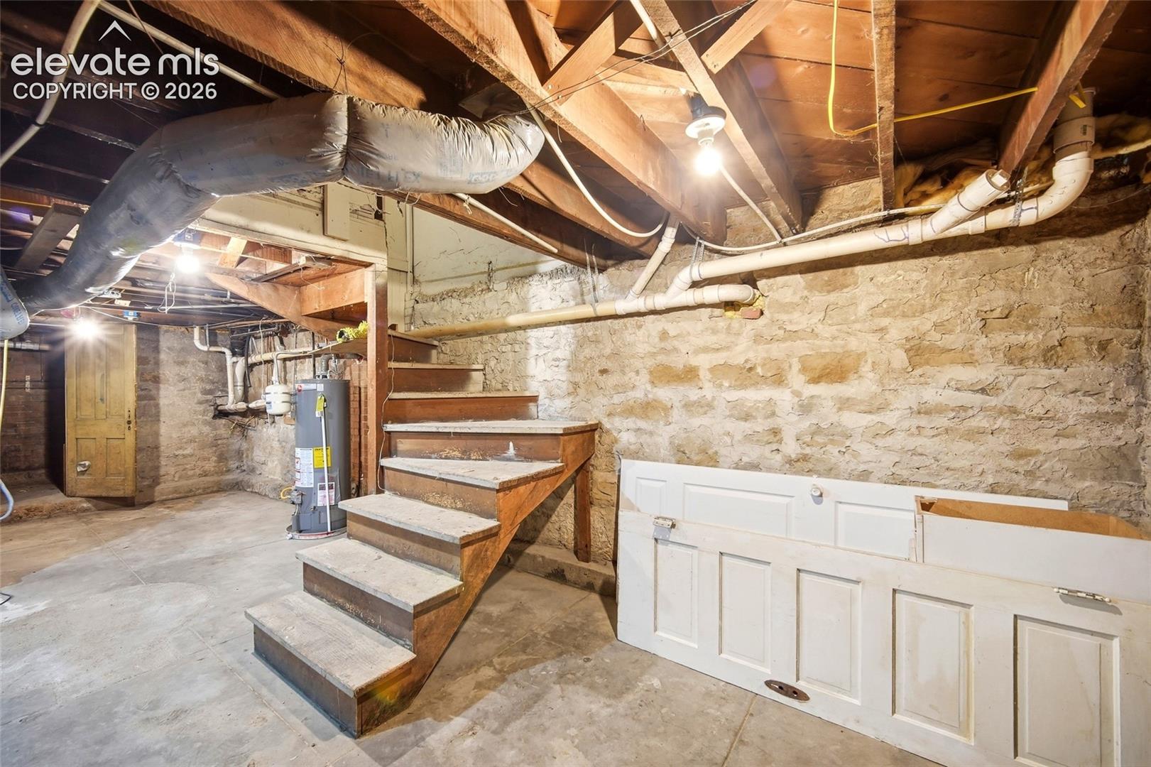 Unfinished basement space featuring exposed wood joist ceilings, stone foundation walls, concrete flooring, and wooden staircase construction