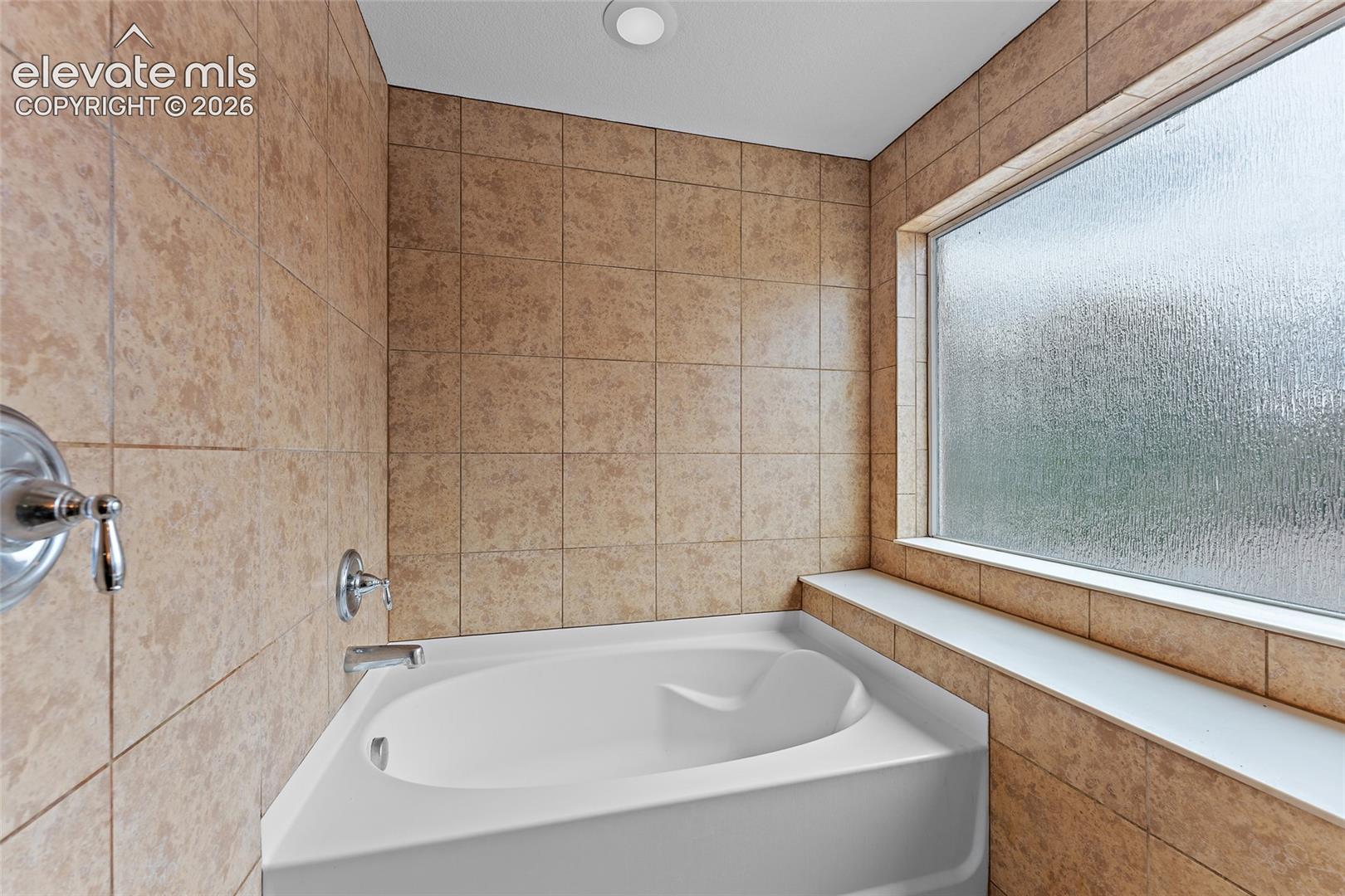 Soaking tub featuring tiled surround, integrated ledge, chrome hardware, and frosted glass window