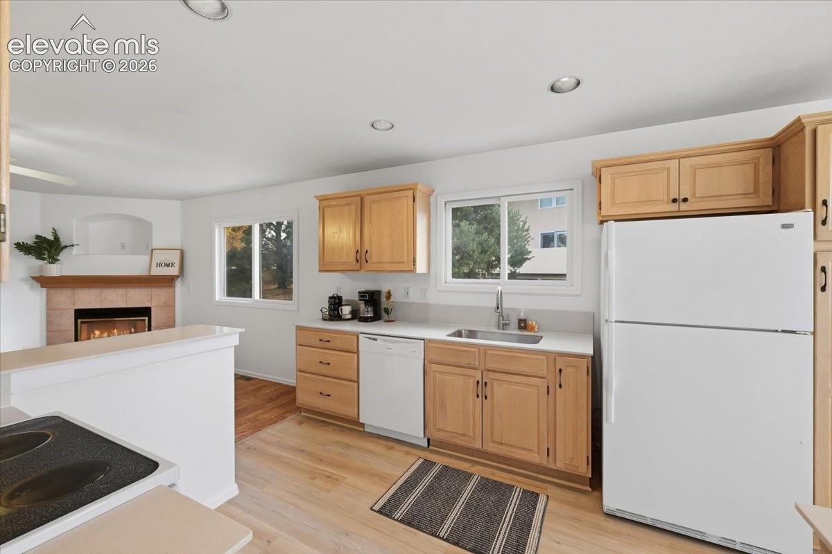 Kitchen featuring light wood cabinetry, white appliances, solid surface countertops, recessed lighting, and wood-finish flooring with a tile-surround fireplace