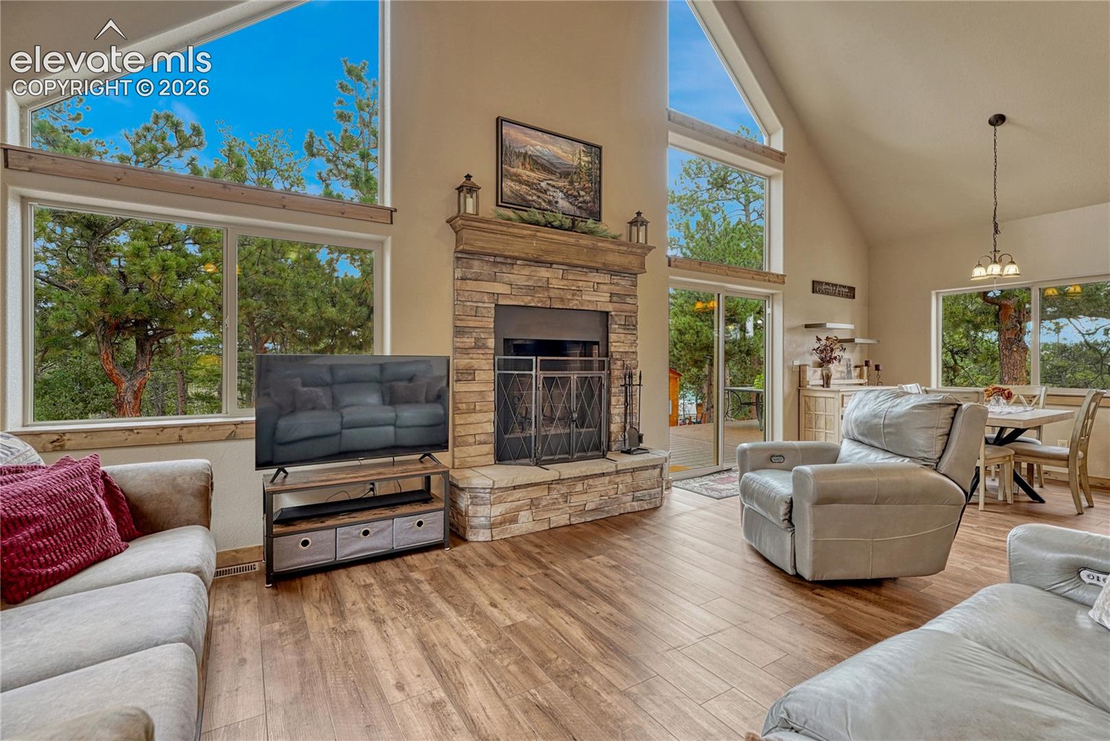 Vaulted ceilings with expansive floor-to-ceiling windows, stone-surround fireplace with a wooden mantel, and wood-finish flooring