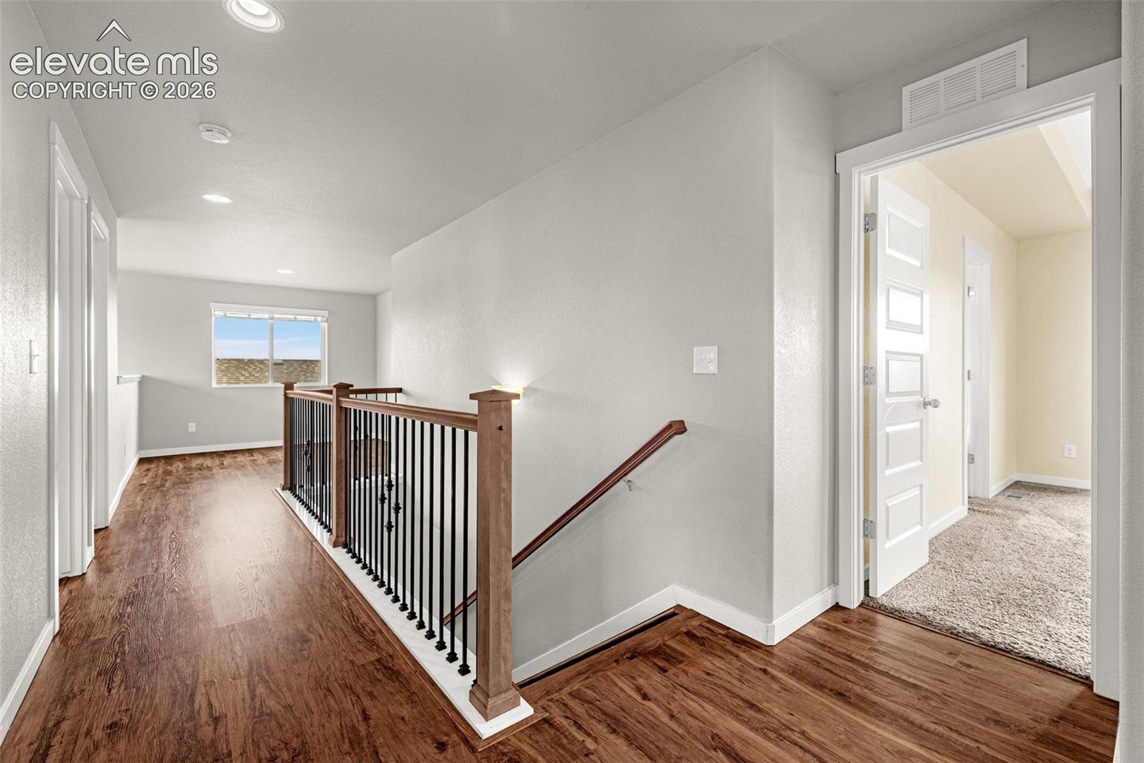 Wood-finish flooring, staircase with dark metal balusters and wood newel posts, recessed lighting, and a white interior door with decorative panels