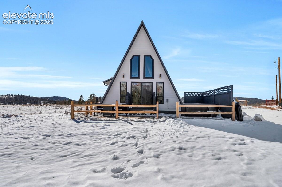 A-frame exterior featuring white horizontal siding, large multi-pane glass windows, exterior wall-mounted lighting, a private fenced perimeter, and a detached privacy screen