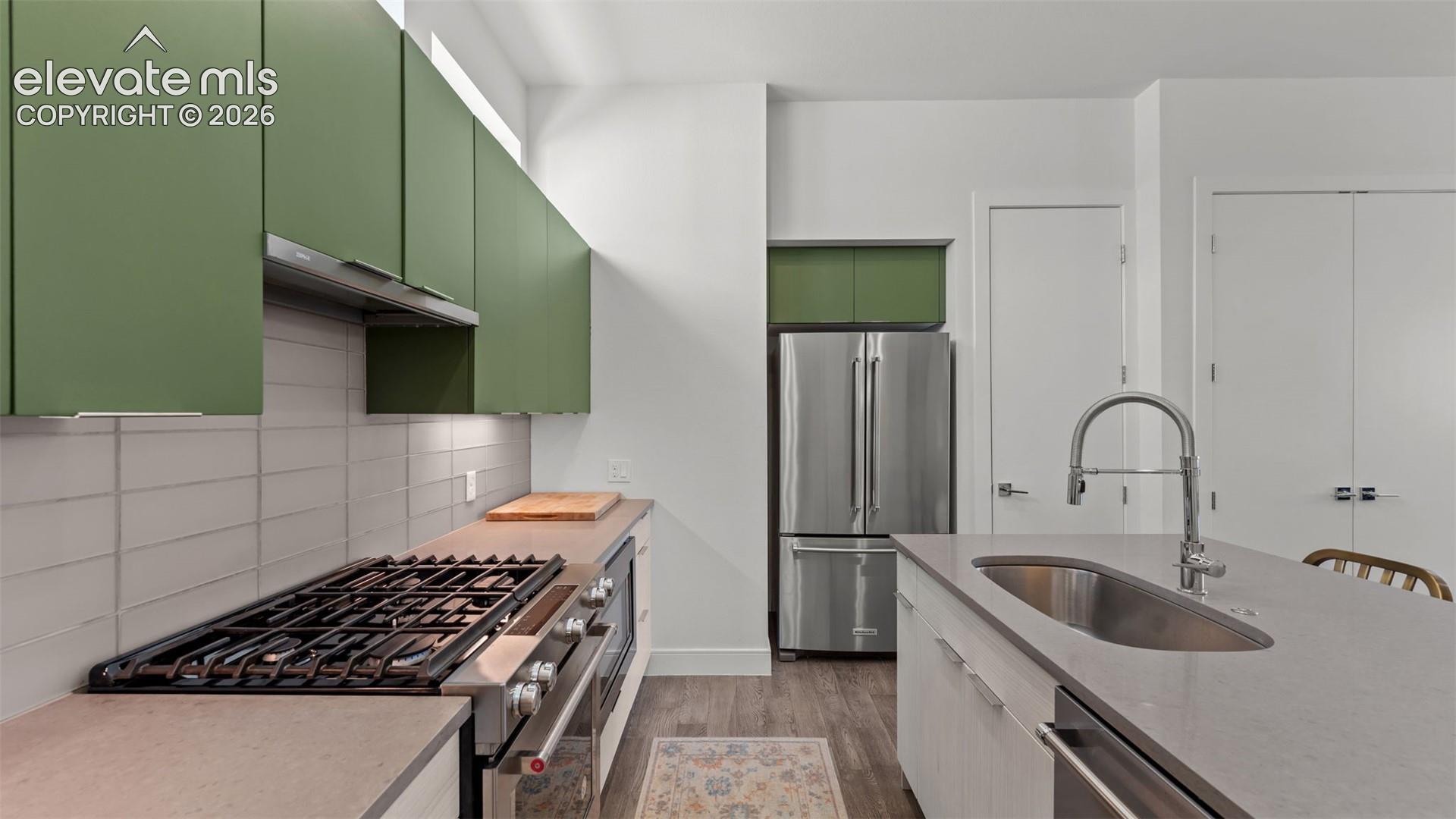 Contemporary kitchen featuring matte green upper cabinetry, stainless steel gas range, light-toned stone-finish countertops, rectangular tile backsplash, and a central island with an undermount sink
