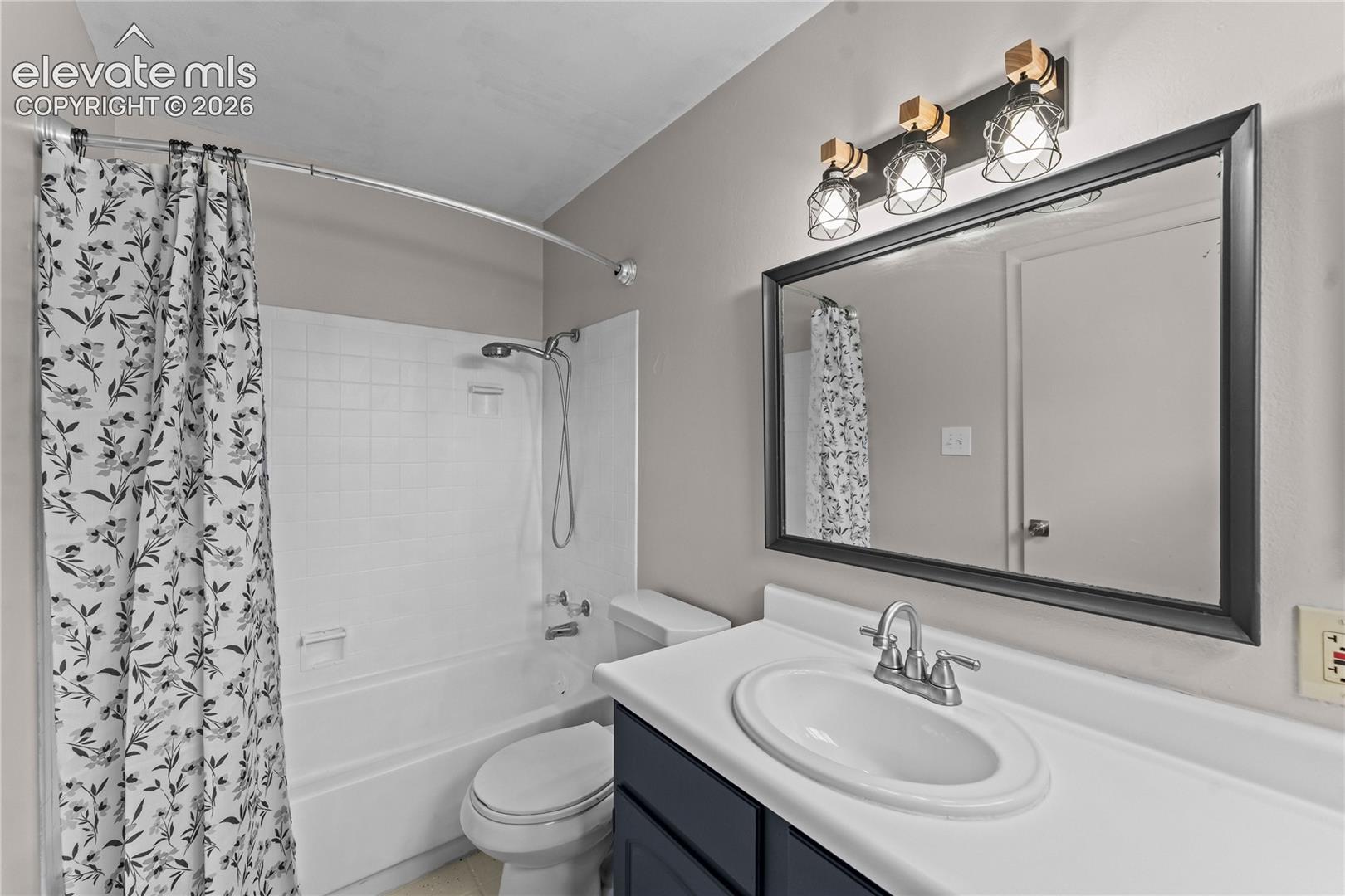 Vanity featuring a dark-toned cabinet base, white integrated sink, three-light decorative fixture, and large framed wall mirror, accompanied by a tiled tub-shower combination