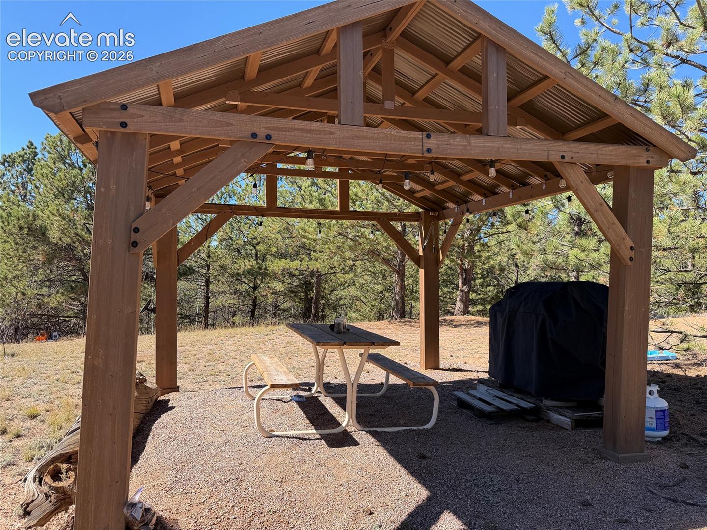 Outdoor timber-frame pavilion featuring a metal roof, exposed wooden trusses, and natural gravel flooring
