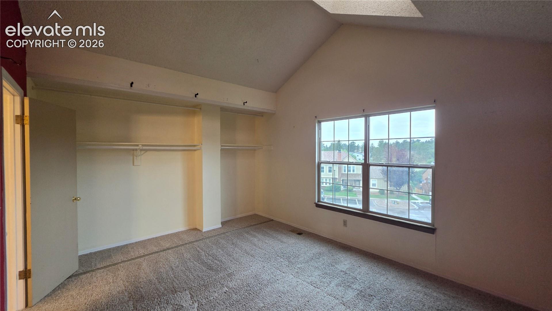 Vaulted ceiling, large window, built-in shelving, carpet flooring, and an open closet system