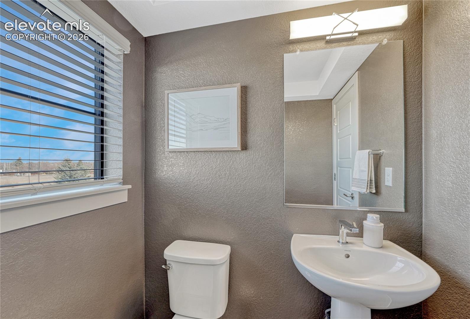 Wall-mounted mirror with integrated vanity lighting, white pedestal sink, polished chrome faucet, and horizontal slat window blinds