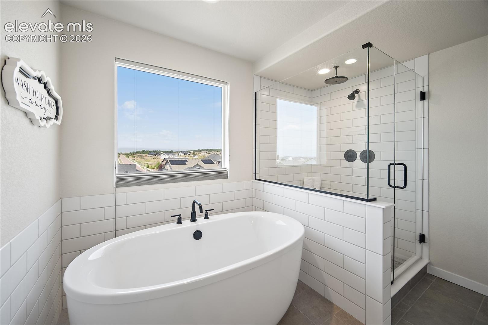 Freestanding soaking tub with matte black fixtures, glass-enclosed shower with subway tile surround, two shower heads, and large window
