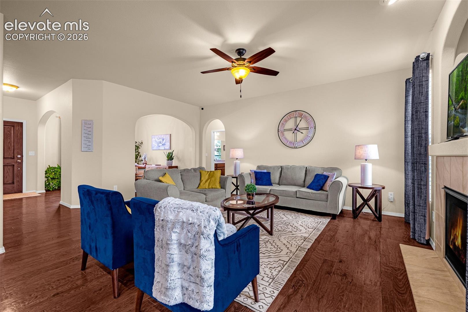 Living area featuring wood-finish flooring, recessed archways, a ceiling fan with light fixture, and a fireplace with tile surround