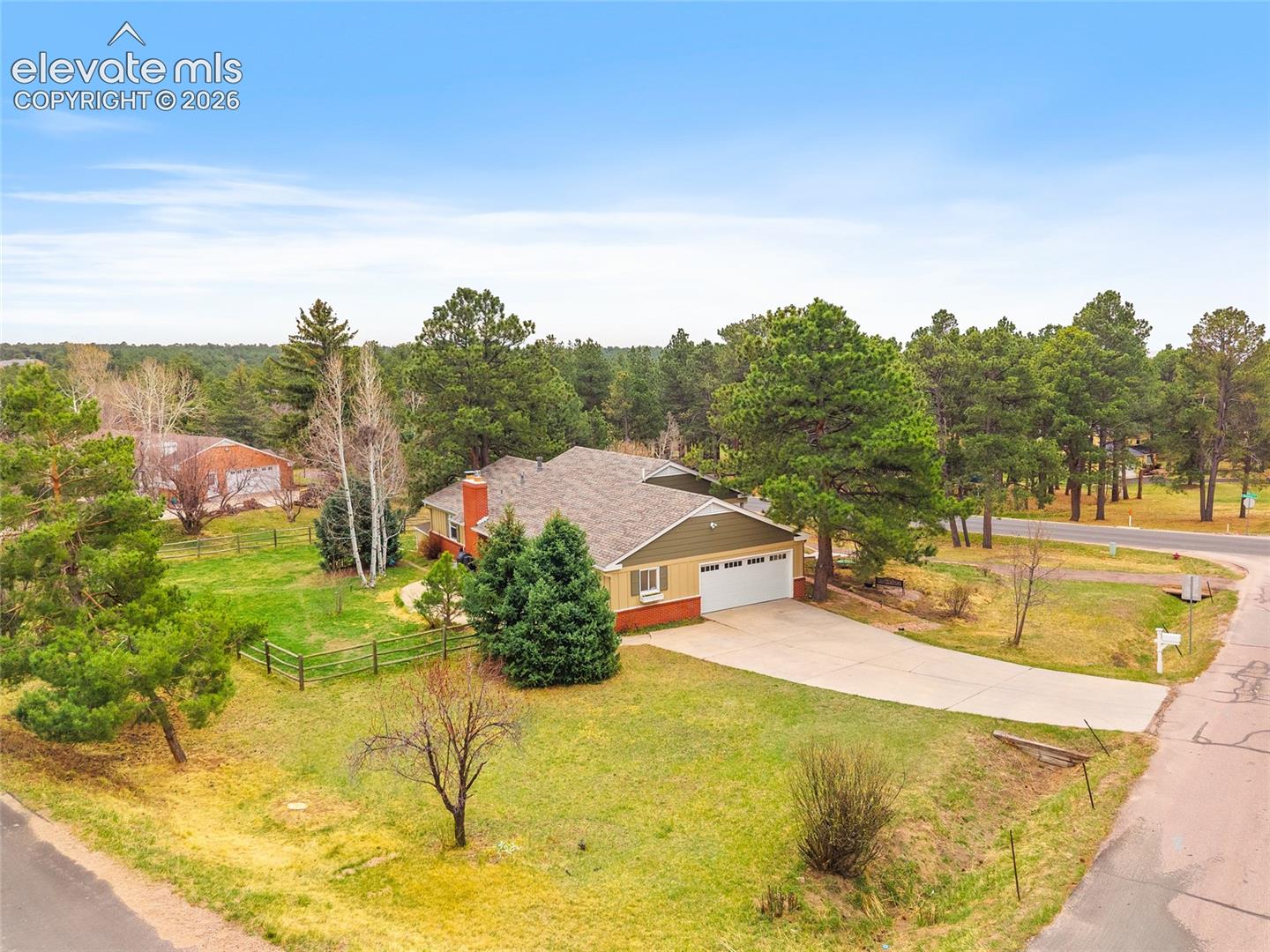 Single-story residence featuring a two-car garage, brick and horizontal siding exterior, attached brick chimney, and concrete driveway situated on a corner lot with mature coniferous trees