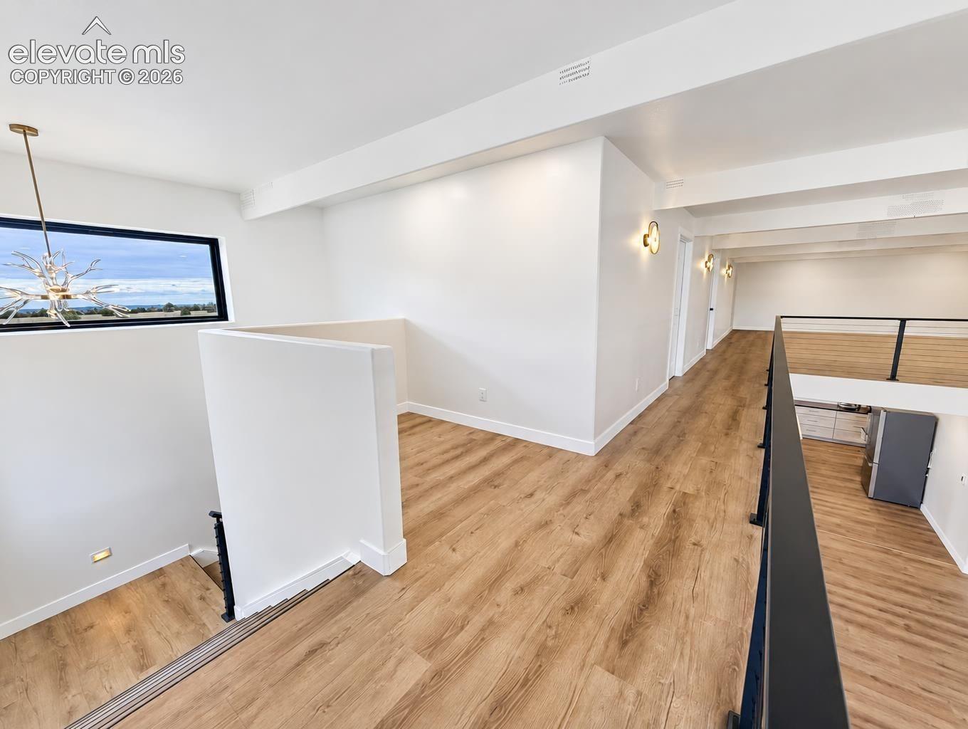 Open floor plan featuring wood-finish flooring, horizontal rectangular window, architectural beam detailing, black metal railing, and wall-mounted light fixtures