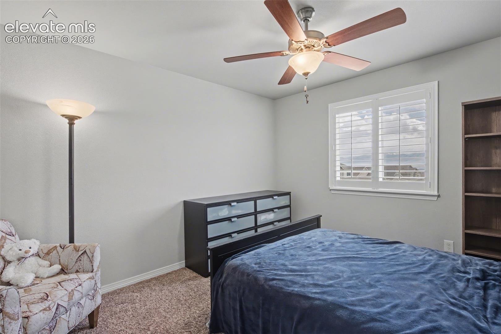 Window with louvered shutters, ceiling fan with integrated lighting, built-in shelving, neutral wall color, and carpeted flooring