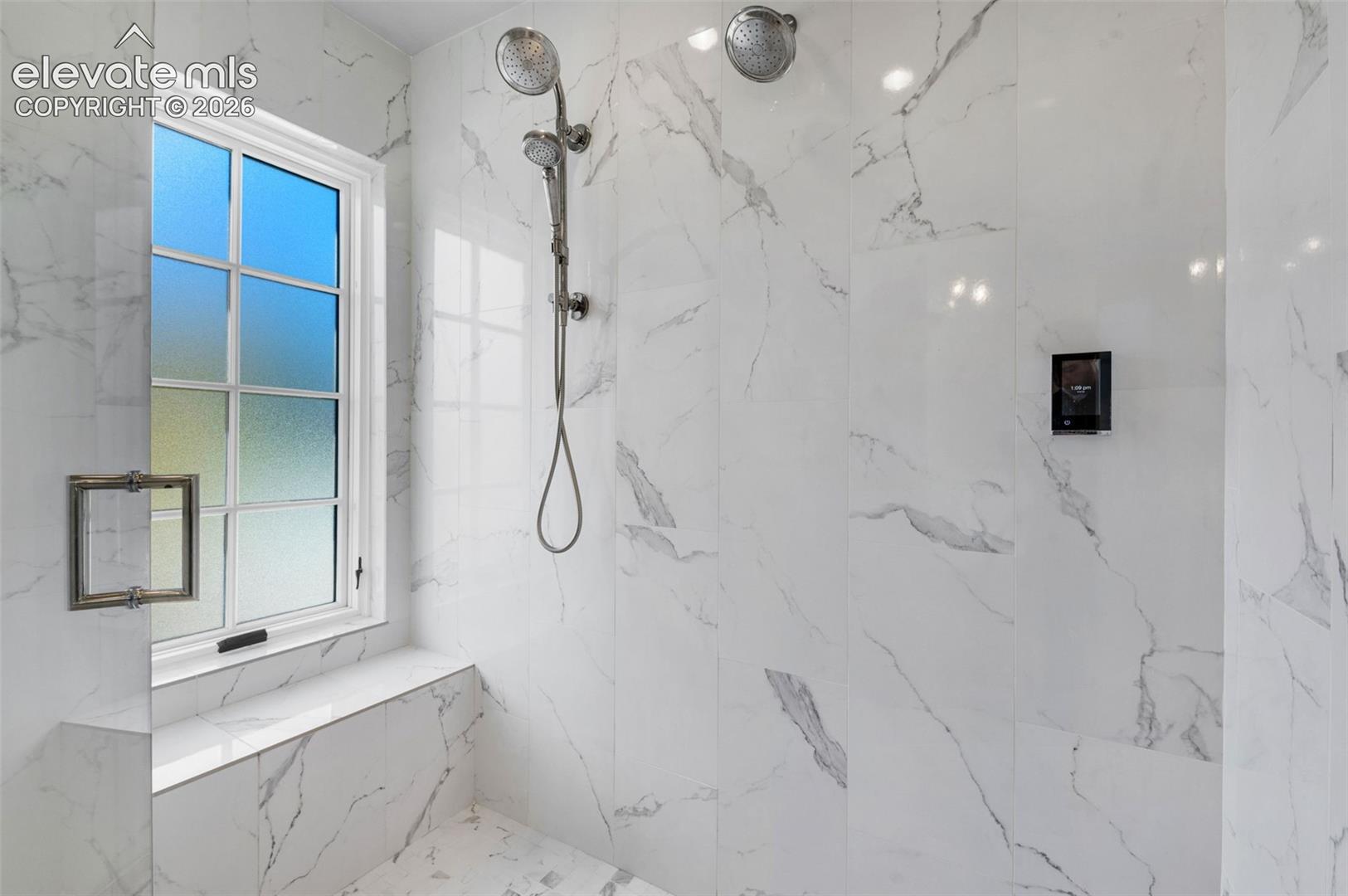 Walk-in shower featuring floor-to-ceiling veined tile, a built-in tiled bench, polished metal dual showerheads, and a privacy-glass window
