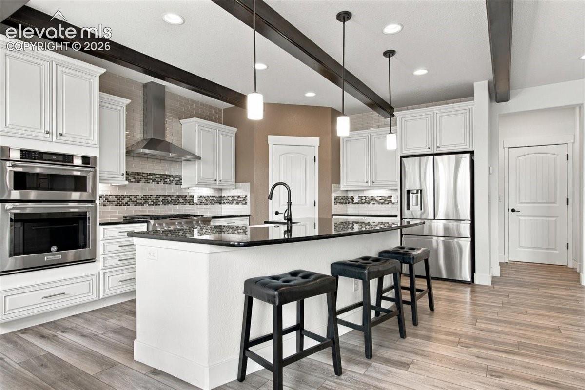 Kitchen featuring white cabinetry, center island with dark stone countertop, built-in stainless steel double wall ovens, wood-finish flooring, and exposed ceiling beams