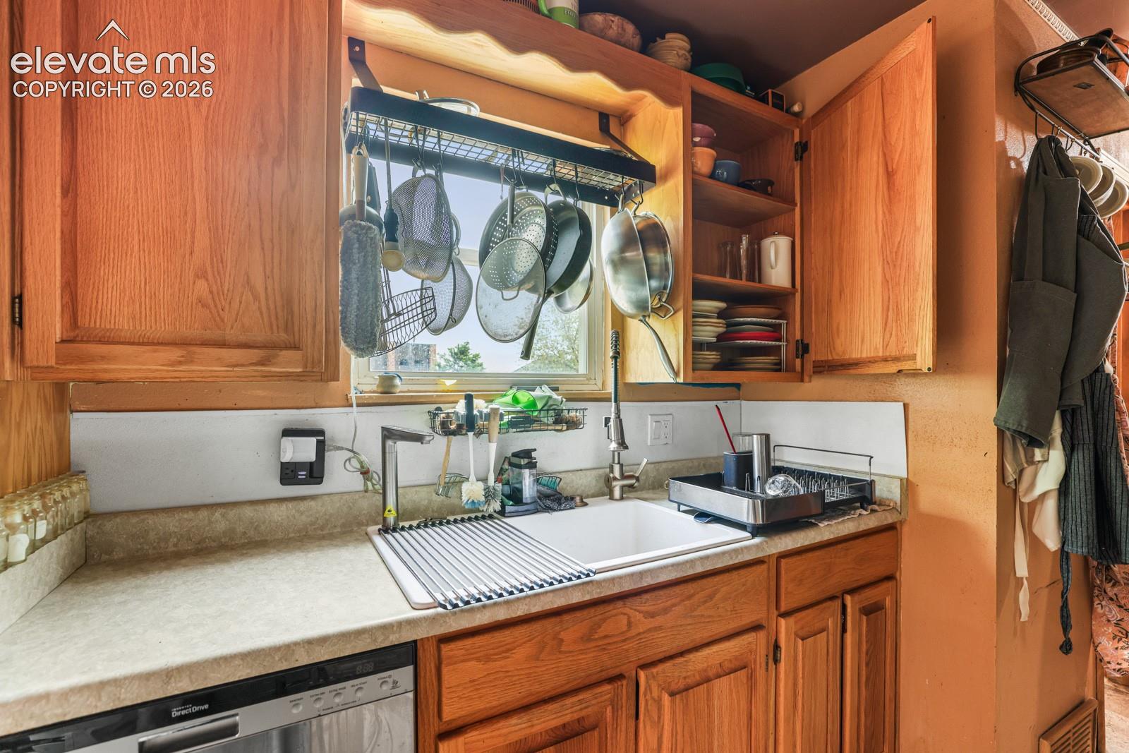 Kitchen workstation featuring light wood cabinetry, a white single-basin sink with a gooseneck faucet, light-toned countertops, a stainless steel dishwasher, and a window with natural light