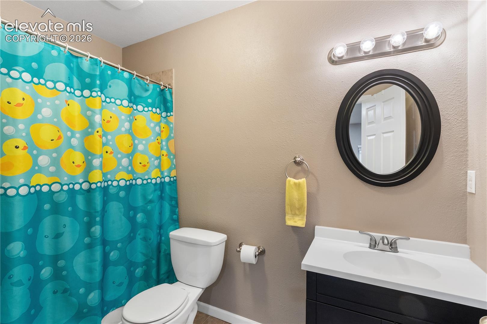 Wall-mounted round mirror with black frame, vanity featuring a white countertop with integrated sink, and four-bulb light fixture