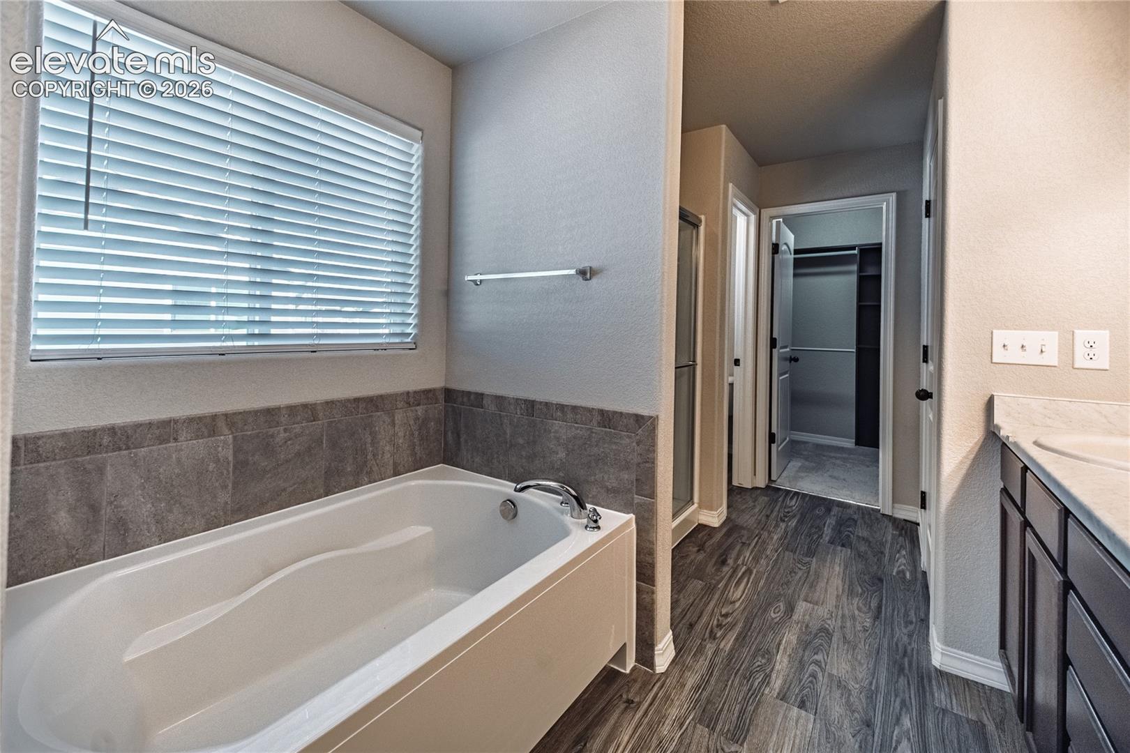 White soaking tub with tiled surround, wood-finish flooring, and a vanity with a light-toned countertop