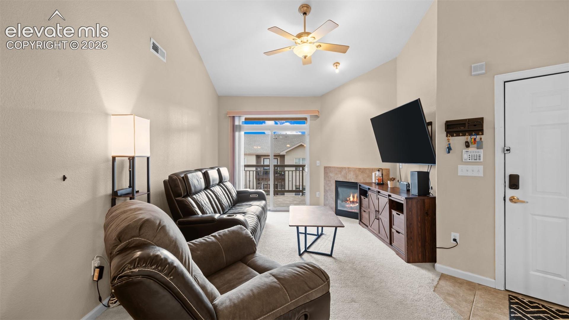 Living area featuring vaulted ceilings, a ceiling fan, a gas fireplace with tile surround, light-toned carpet flooring, and a sliding glass door leading to an outdoor balcony