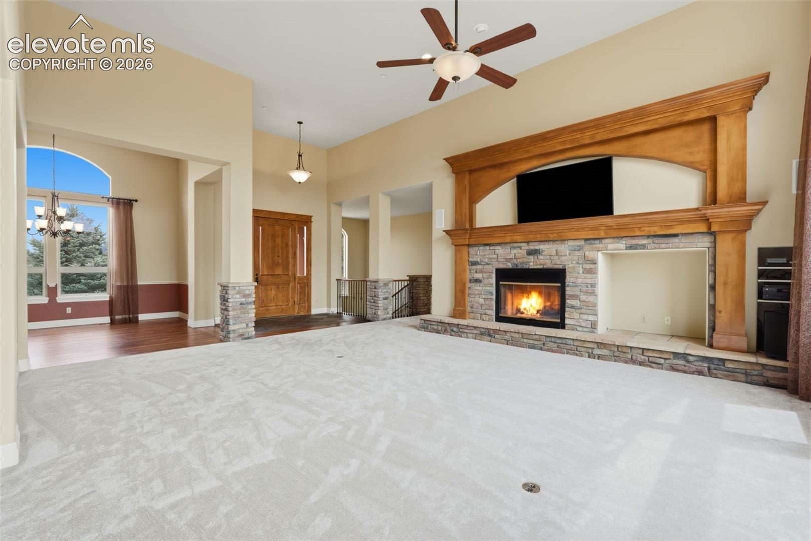 Spacious living area featuring a stone-clad fireplace with a prominent wood mantel, high ceilings, and light-toned carpeting
