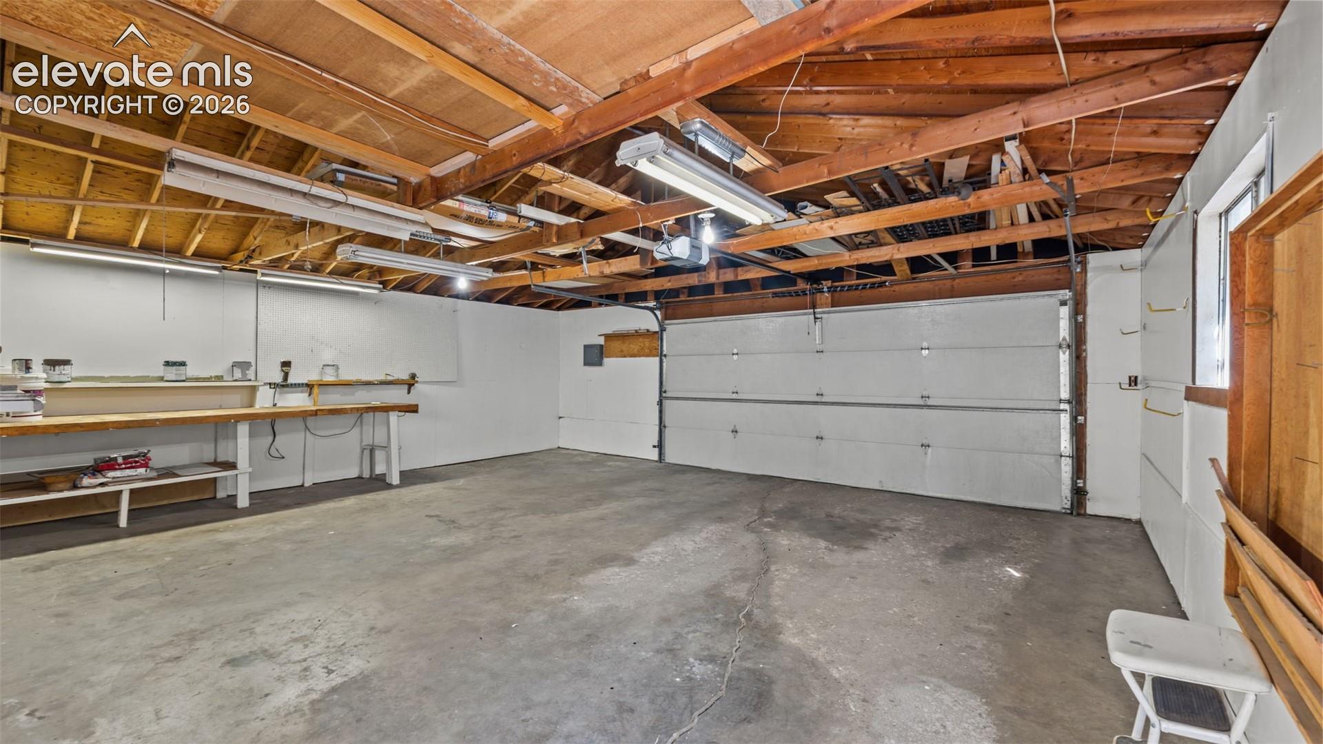 Concrete flooring garage with an exposed wood beam ceiling, white walls, built-in workbench, and overhead fluorescent lighting