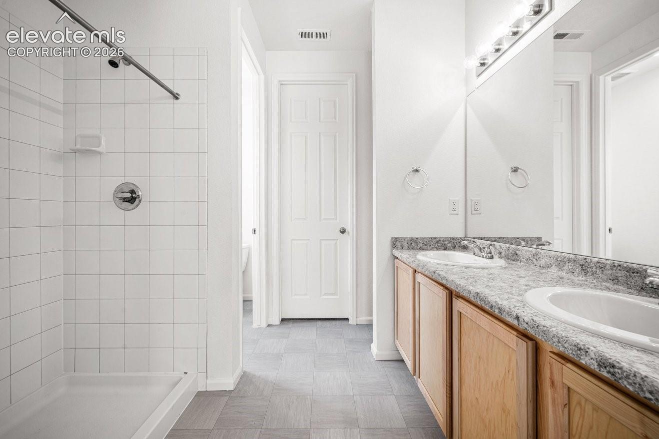 Spa-inspired primary bathroom featuring a spacious walk-in shower with tile surround and glass enclosure, dual-sink vanity with wood-finish cabinetry and stone countertops, large mirror with integrated lighting, and neutral tile flooring throughout